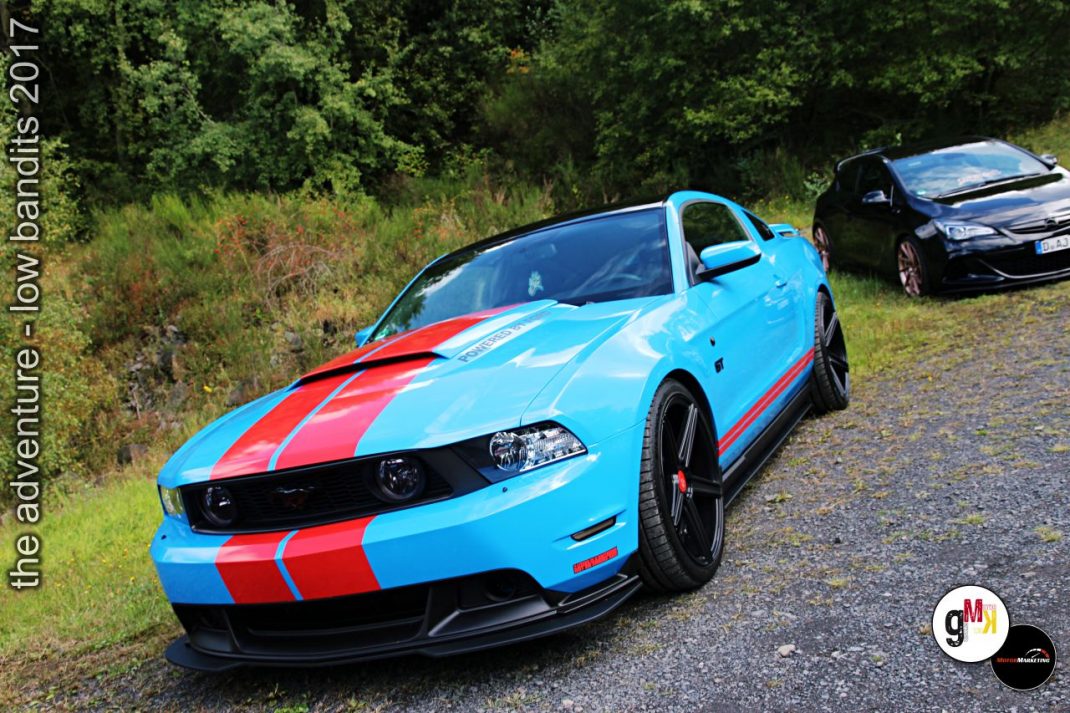 Hellblauer Muscle-Car mit roten Streifen und schwarzem Youngtimer im Hintergrund auf Waldweg bei Tuning-Veranstaltung