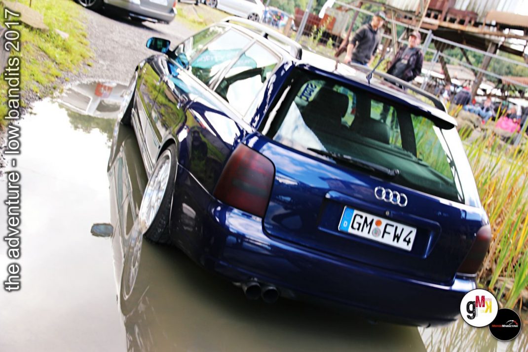 Blauer Audi-Youngtimer mit offener Tür fährt durch Wasserpfütze auf unbefestigtem Gelände beim Tuning-Event