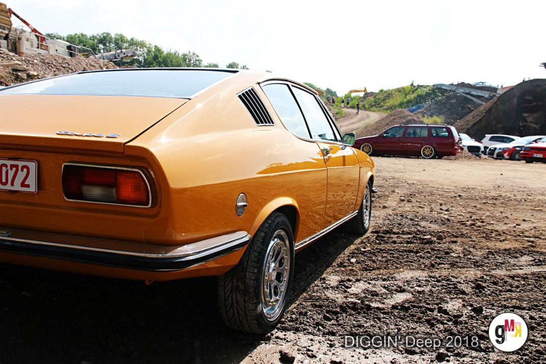 Orangefarbener Oldtimer-Coupé in Seitenansicht auf Schotterfläche, weitere Fahrzeuge im Hintergrund bei der DIGGIN' Deep 2018 Rallye in der Eifel