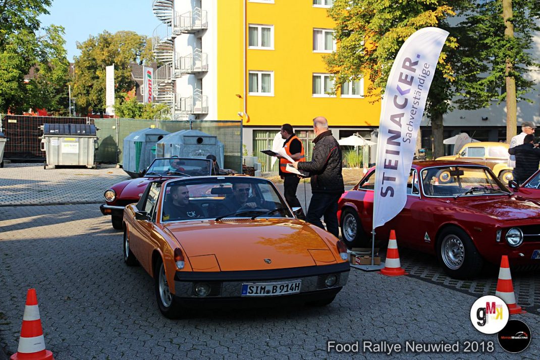 Orangefarbener Sportwagen und weitere Oldtimer auf Parkplatz bei der Food Rallye Neuwied, Organisatoren kontrollieren Teilnehmer, gelbes Gebäude im Hintergrund
