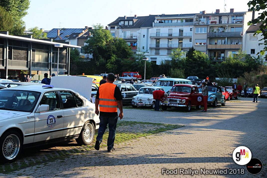 Oldtimer-Rallye-Teilnehmer in orangefarbener Weste kontrolliert Fahrzeugkolonne auf Parkplatz in Neuwied, Zuschauer und Helfer zwischen geparkten Klassikern