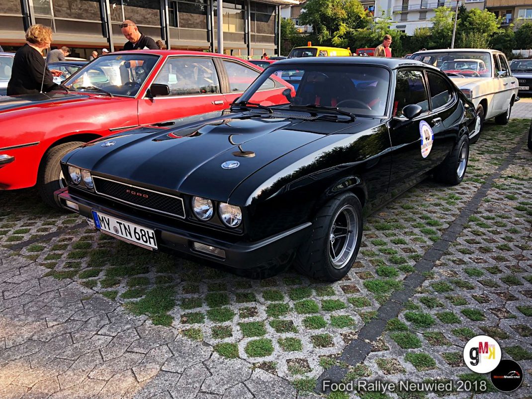 Schwarzer Oldtimer-Sportwagen mit Motorhaube-Streifen auf Pflasterfläche, daneben rote Cabrios bei der Food Rallye Neuwied