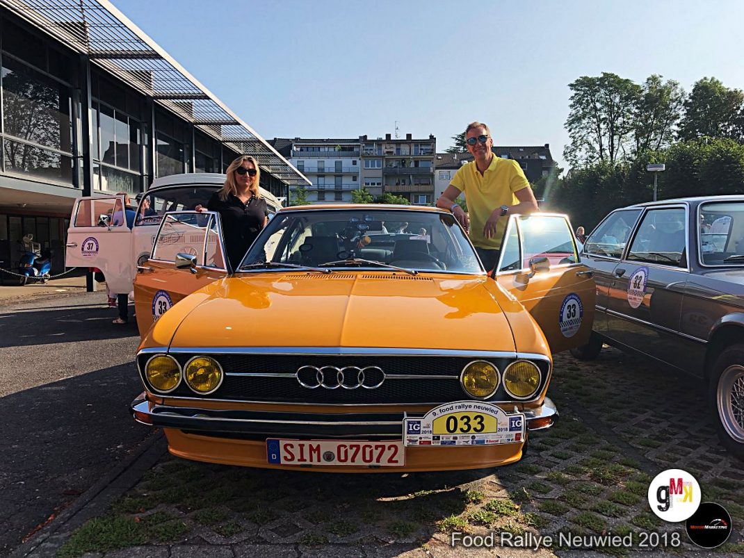 Orange Audi Oldtimer mit Rallye-Startnummern auf Parkplatz, Fahrer und Beifahrerin posieren, weitere Fahrzeuge im Hintergrund, Food Rallye Neuwied 2018