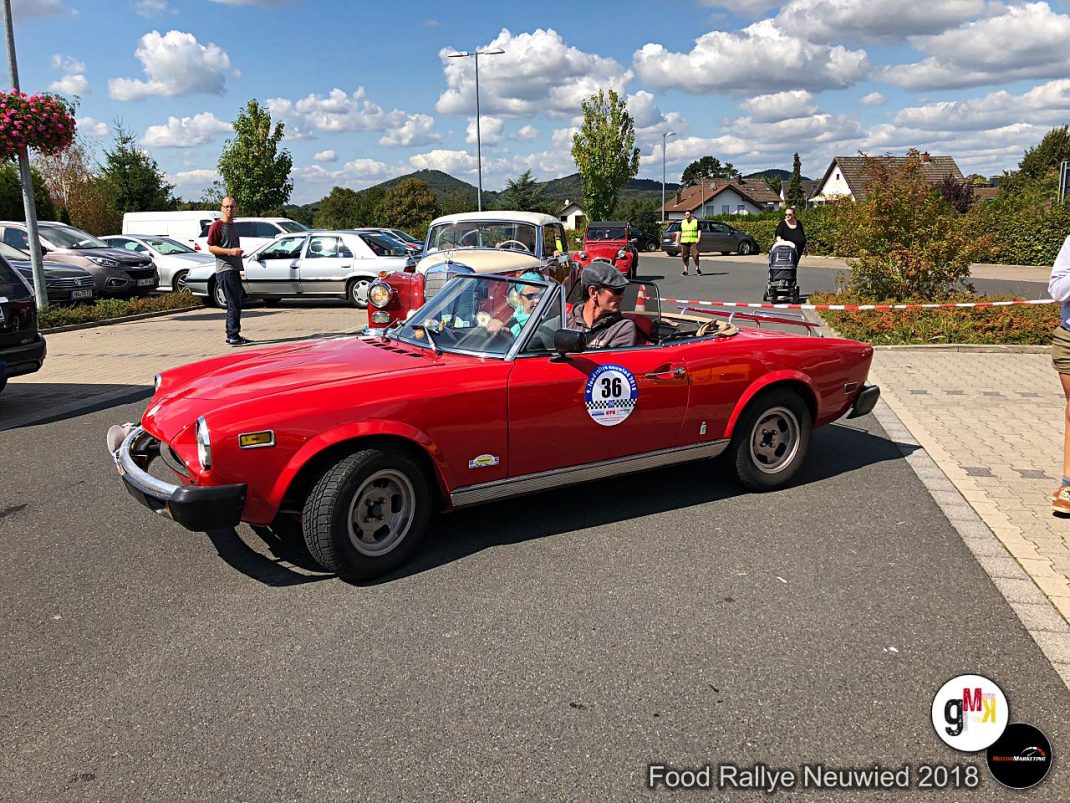 Rotes Oldtimer-Cabrio mit Rallyenummern bei der Food Rallye Neuwied 2018, Fahrer und Beifahrer im Fahrzeug, Parkplatz mit weiteren Fahrzeugen im Hintergrund