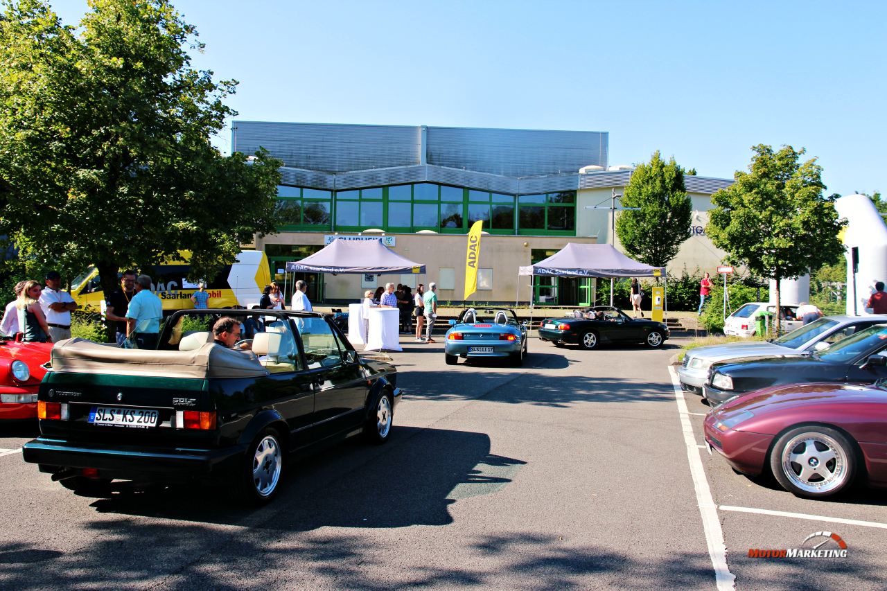 Teilnehmer verschiedener Youngtimer-Fahrzeuge versammeln sich auf einem Parkplatz vor Gebäuden zur ADAC Youngtimer Tour Saarland