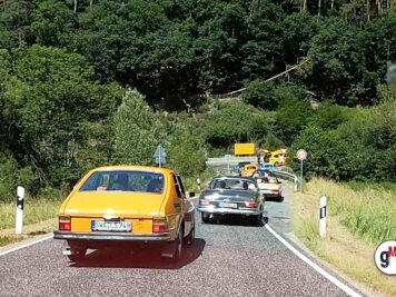 Gelber Oldtimer-Sedan und dunkler Sportwagen fahren auf Landstraße durch bewaldete Hügellandschaft bei ADAC Oldtimerfahrt Hessen-Thüringen