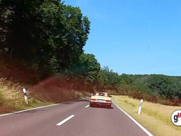 Oldtimer-Cabrio fährt auf gerader Landstraße mit bewaldeten Hügeln und rötlichen Böschungen während der ADAC Oldtimerfahrt Hessen-Thüringen
