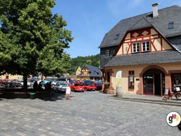 Oldtimer-Rallye auf Dorfplatz mit Fachwerk-Gebäuden und geparkten klassischen Automobilen verschiedener Epochen