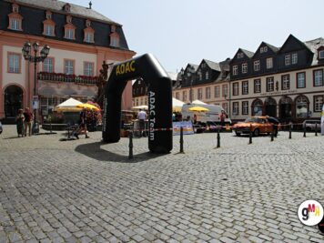 ADAC Oldtimerfahrt auf historischem Marktplatz mit schwarzem Startbogen, Oldtimern und Zuschauern unter Sonnenschirmen