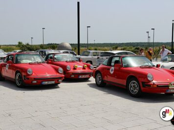 Mehrere rote Porsche-Oldtimer stehen auf einem Parkplatz bei der ADAC Oldtimerfahrt Hessen-Thüringen, Teilnehmer im Hintergrund