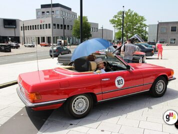 Roter Oldtimer-Cabrio mit Startnummer auf Parkplatz, Fahrer mit blauem Sonnenschirm, weitere Fahrzeuge und Teilnehmer im Hintergrund bei der ADAC Oldtimerfahrt
