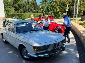Silberner Oldtimer-Klassiker parkt auf Veranstaltungsgelände, Teilnehmer in heller Kleidung stehen bei roten Youngtimern im Hintergrund