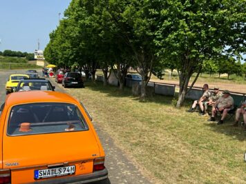 Orange Fiat 126 in einer Kolonne von Oldtimern bei einer Fahrt unter schattenspendenden Bäumen, Teilnehmer ruhen sich aus