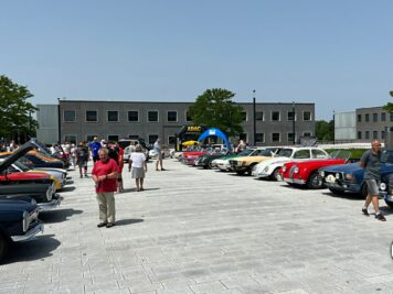 Oldtimer verschiedener Epochen auf großem Platz mit Besuchern bei ADAC Oldtimerfahrt, Industriegebäude im Hintergrund