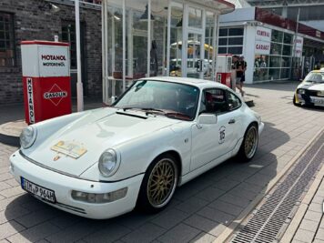 Weißer Porsche 911 mit Startnummer an einer Rallyestation mit historischem Gaskutschen-Schild und anderen Oldtimern