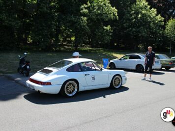 Weißer Porsche 911 Carrera mit Startnummer 115 bei der ADAC Oldtimerfahrt Hessen-Thüringen, umgeben von Teilnehmern und Fahrzeugen in grüner Parklandschaft