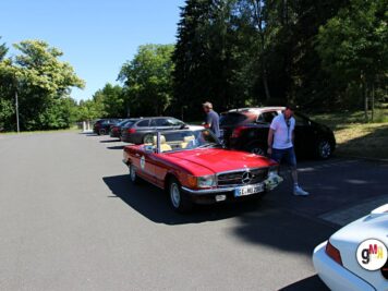 Rotes Oldtimer-Cabrio parkt zwischen anderen Fahrzeugen auf Parkplatz, Teilnehmer stehen daneben, Bäume im Hintergrund