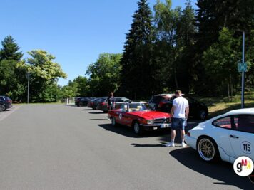 Oldtimer-Cabrio in Rot und Sportwagen in Weiß mit Startnummer auf Parkplatz während ADAC Oldtimerfahrt, Teilnehmer zwischen Fahrzeugen, Bäume im Hintergrund