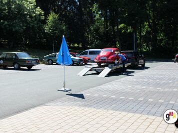Mehrere Oldtimer parken auf gepflastertem Platz unter Bäumen, ein rotes Fahrzeug steht auf Transportanhänger zur ADAC-Oldtimerfahrt