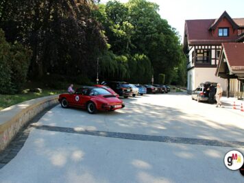 Roter Oldtimer-Sportwagen mit Startnummer fährt in Parkplatz-Gelände ein, weitere Fahrzeuge und Fachwerkhaus im Hintergrund