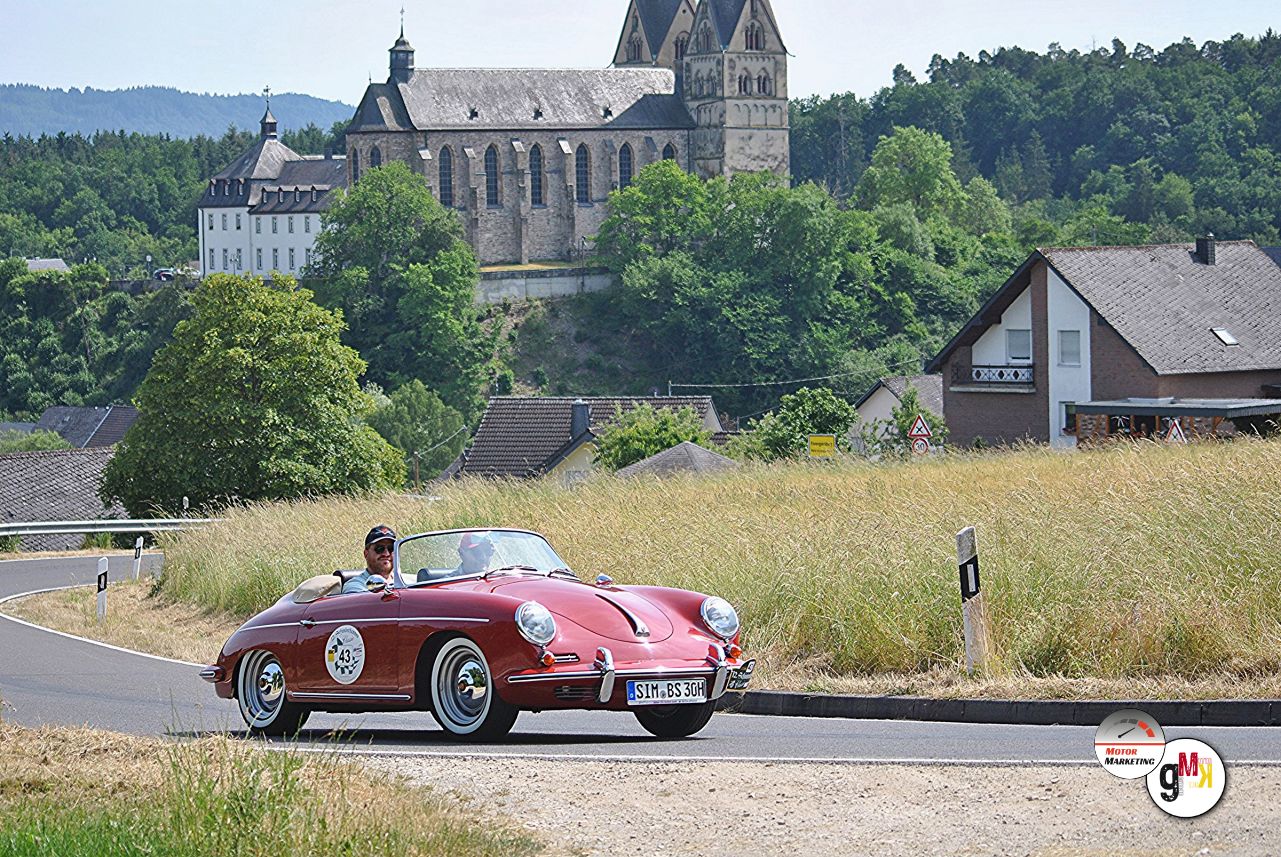 Roter Oldtimer-Sportwagen auf kurvenreicher Landstraße vor Kirchenkulisse und Wäldern während der Hunsrücker Herbst-Classic