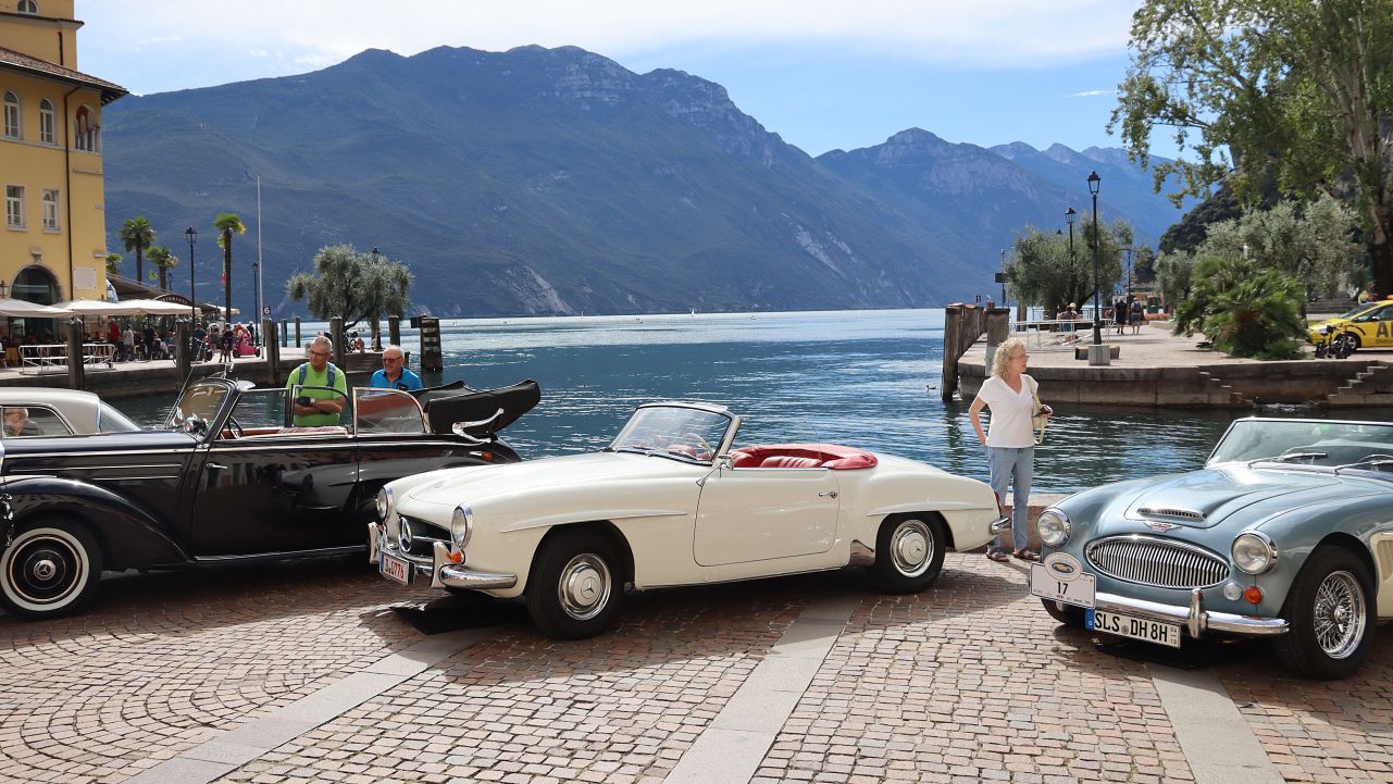 Drei Oldtimer-Cabriolets stehen an der Uferpromenade des Gardasees, im Hintergrund Berge und ein gelbes Gebäude