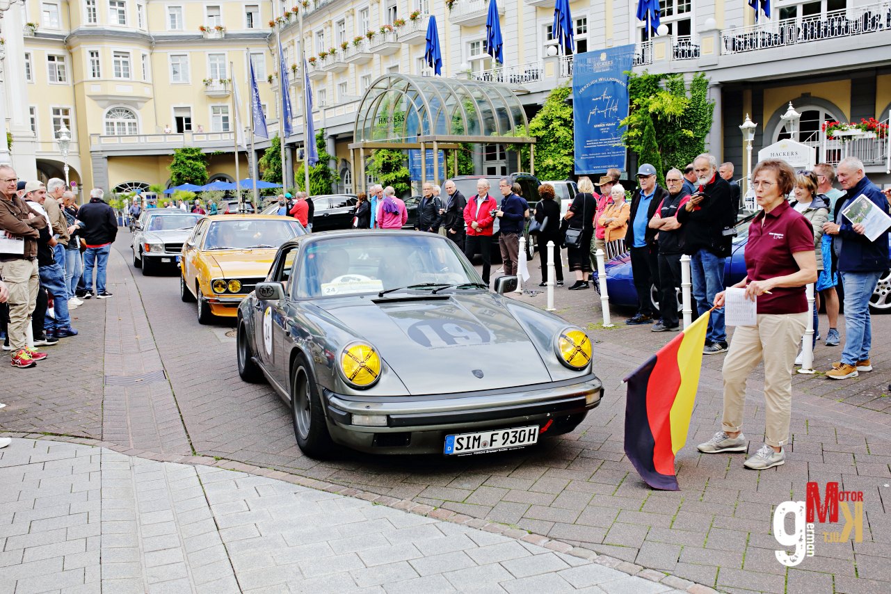 Porsche 911 mit gelben Scheinwerfern in Hotelinnenhof, umgeben von Teilnehmern und deutschen Flaggen der ADAC Mittelrhein Classic Rallye