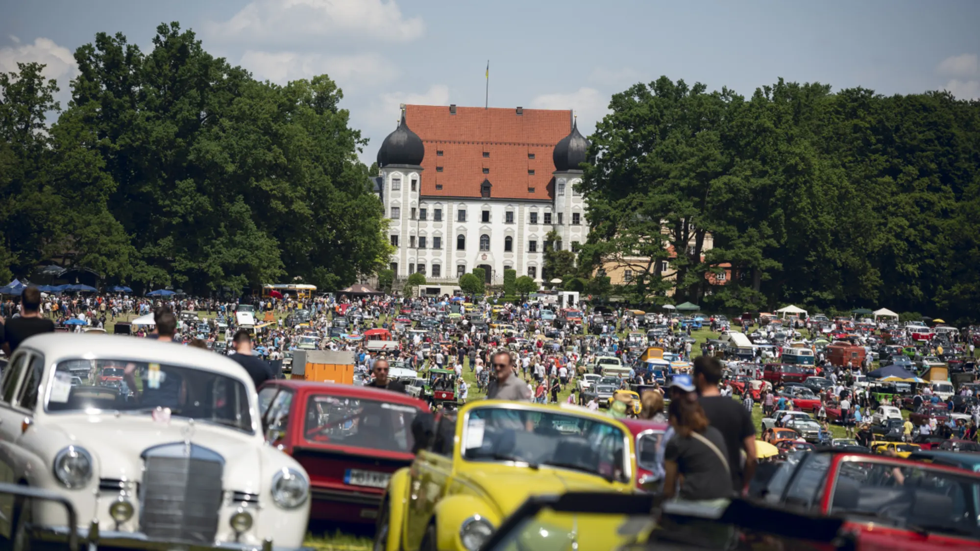 Oldtimer-Cabrios und Limousinen aus verschiedenen Jahrzehnten auf einem Schlossgelände mit Zuschauermenge und Schloss mit roten Dächern im Hintergrund