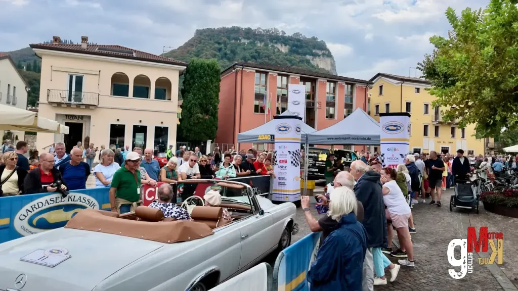 Oldtimer-Cabrio auf Marktplatz bei Gardasee-Klassik-Veranstaltung mit Besuchern, Zelten und italienischer Bergkulisse