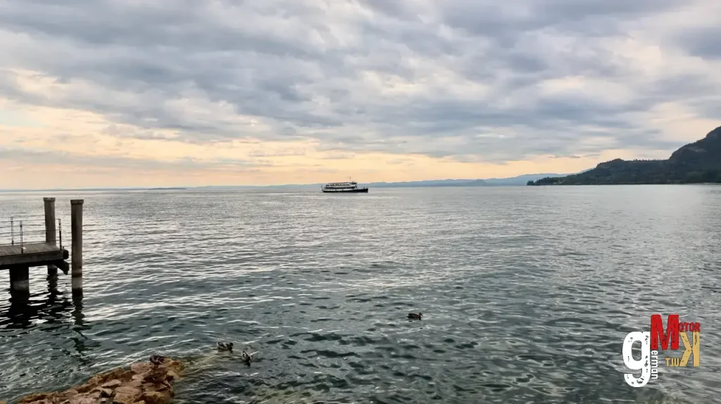 Holzsteg und Uferblick auf den Gardasee mit Fähre und bewaldeter Bergkulisse zur ADAC Gardasee Klassik