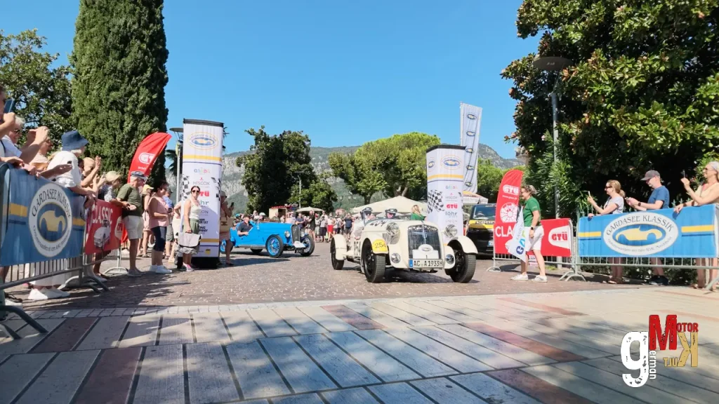 Oldtimer-Rennwagen fahren beim Start einer Rallye an Zuschauern und Bannern vorbei, im Hintergrund Berge und mediterrane Vegetation