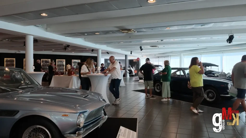 Teilnehmer und Besucher in einer Ausstellungshalle mit mehreren Oldtimern und Informationsständen zur ADAC Gardasee Klassik