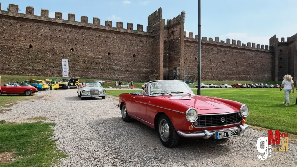 Rotes Oldtimer-Cabrio vor mittelalterlicher Burgmauer bei der ADAC Gardasee Klassik, weitere Youngtimer auf Rasenplatz