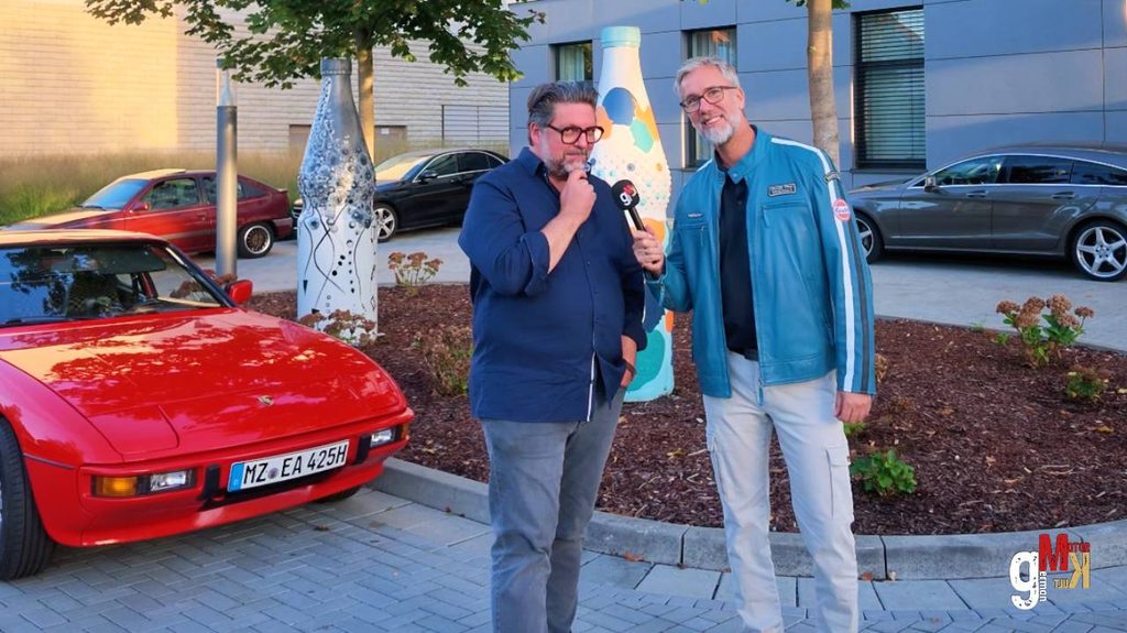 Zwei Moderatoren mit Mikrofonen vor einem roten Sportwagen und weiteren Fahrzeugen bei einer Veranstaltungspräsentation