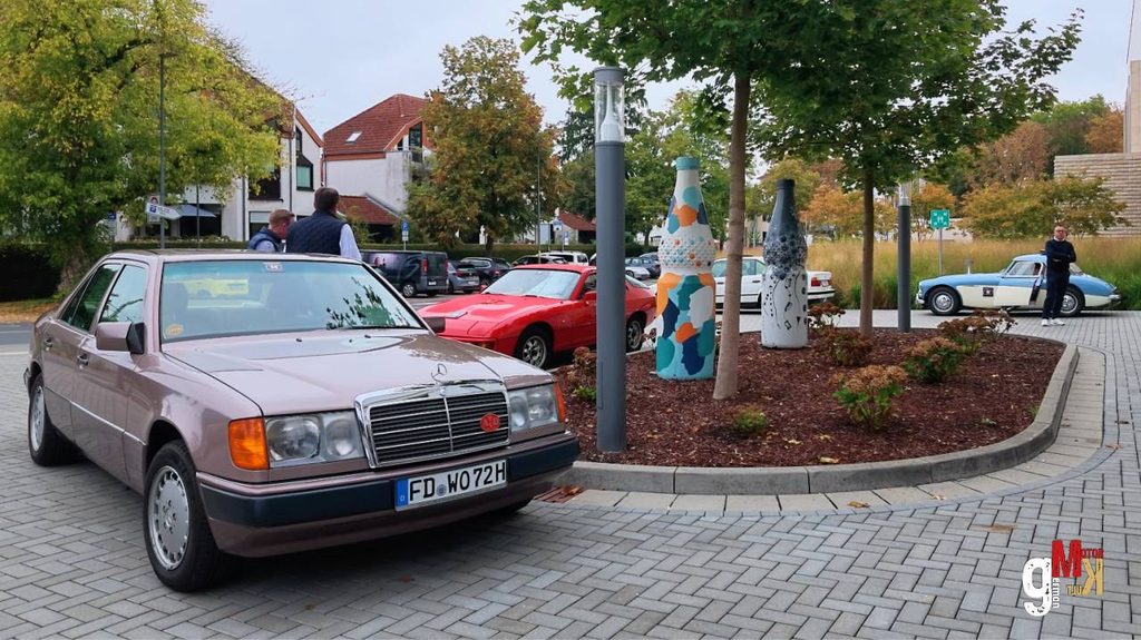 Silberner Oldtimer-Pkw der 1980er Jahre auf gepflastertem Parkplatz bei Rallye-Veranstaltung, im Hintergrund weitere Fahrzeuge und bemalter Straßenbaum
